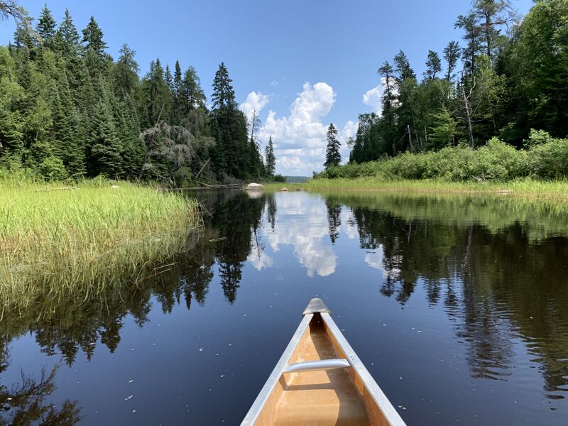 The image shows a canoe gliding through a calm river, flanked by lush green trees and vegetation on both sides. The water reflects the sky and trees, creating a mirror-like effect. The sky is blue with scattered white clouds. The scene conveys a sense of tranquility and immersion in nature.